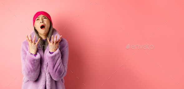 Miserable asian senior woman in stylish purple faux fur coat, pleading god and asking why, shaking