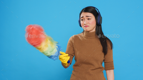 Exhausted sleepy housekeeper wearing headphones while doing cleaning in house using feather duster