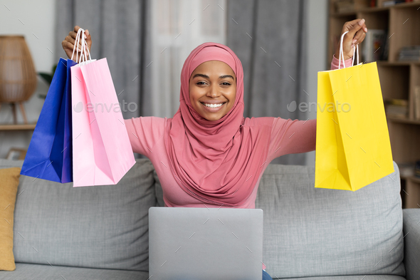 Excited black lady in hijab sitting on sofa with laptop and lifting gift bags above her head, happy
