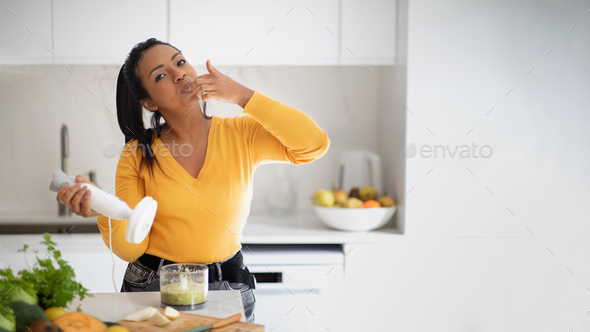 Cheerful millennial african american lady make smoothie with blender, licking finger at table with