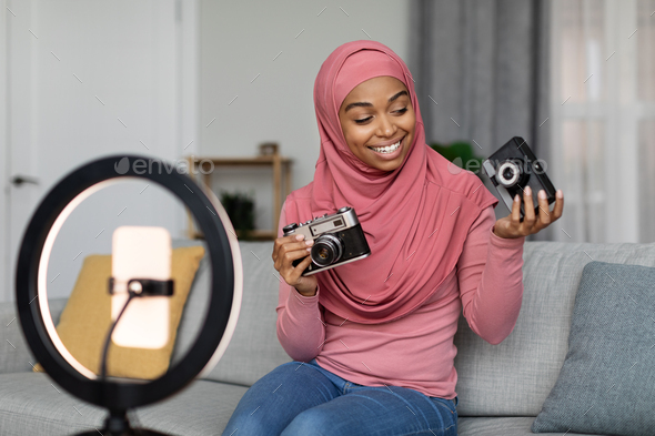 Excited black muslim lady shooting video for blog, making review on retro photo cameras, sitting at