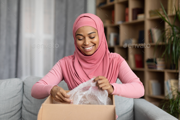 Fast delivery concept. Happy black lady in hijab received package, unpacking cardboard box, sitting