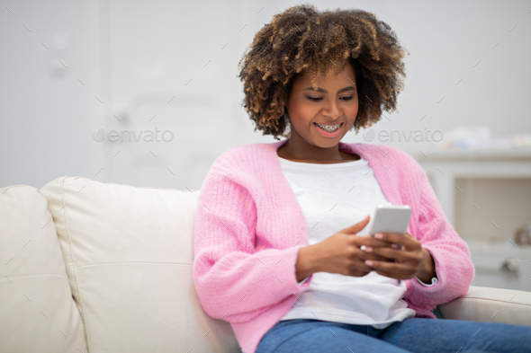 Happy black lady sitting on couch with smartphone, copy space