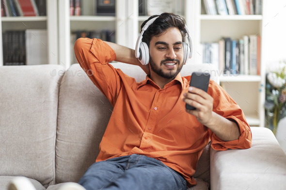 Positive arab guy chilling on couch at home, watching video