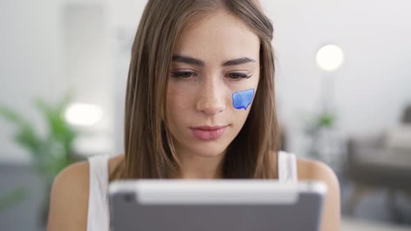 Close-up Portrait of Young Cute Woman Sitting at Home, Looking in the Tablet and Smiling alt