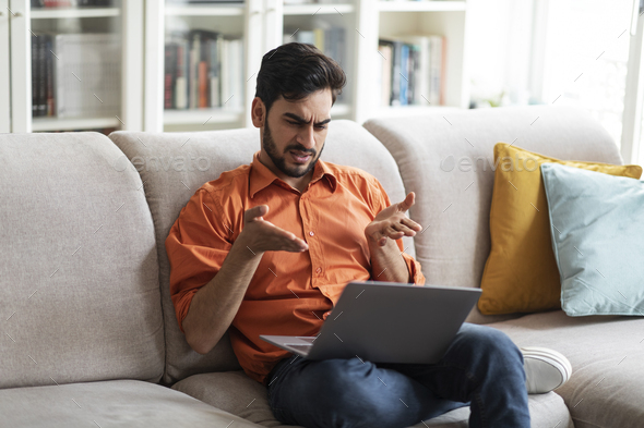 Irritated middle eastern man sitting on couch, using laptop