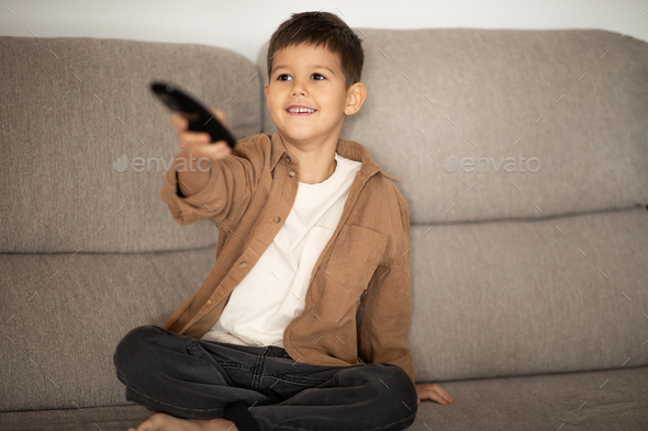 Cheerful caucasian little child with remote control switches channels on TV set, chooses cartoon