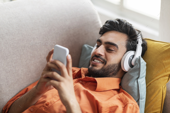 Happy arab guy chilling on couch at home, listening music