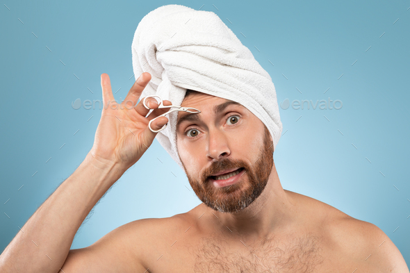 Hair removal concept. Funny bearded man using tweezers for eyebrows, standing with towel on head