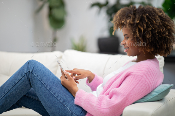 Side view of black woman chilling on couch, using phone