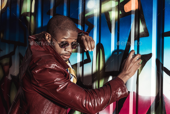 Handsome african american man in stylish clothes painting graffiti with spray paint and looking at