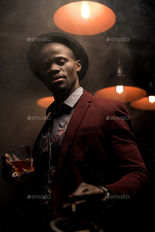handsome luxury african american man in hat smoking cigar and drinking whiskey in smoky room with