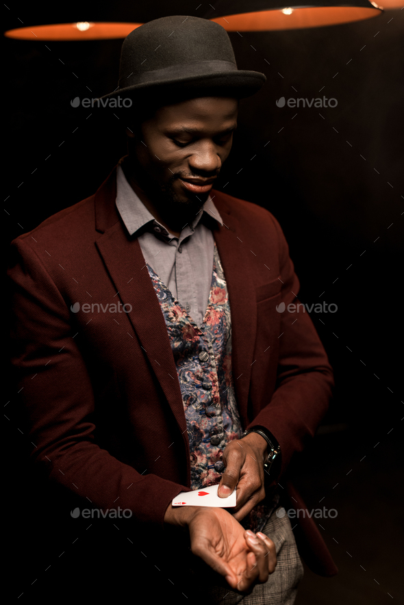 handsome elegant african american man with ace in sleeve in dark room with lamps