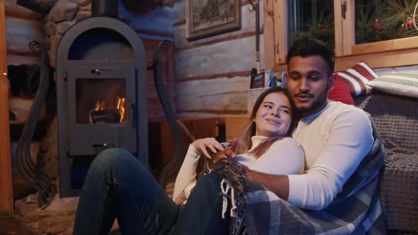 Happy Young Multiracial Couple Relaxing Near the Fireplace Covered with Blanket alt