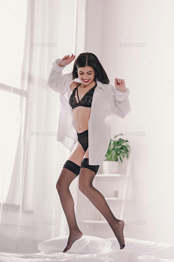 happy attractive girl in black sexy lingerie and stockings jumping on bed  Stock Photo by LightFieldStudios