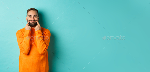 Image of bearded man stretching lips in happy smile, faking happiness, standing over light blue