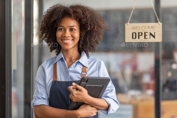 Successful african woman in apron standing coffee shop door. Happy small business owner holding tab