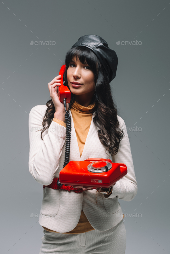 attractive brunette woman in white suit talking by red landline phone isolated on gray