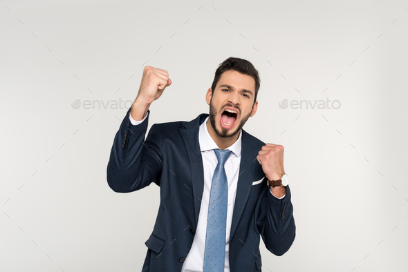 excited young businessman shaking fists and screaming at camera isolated on grey