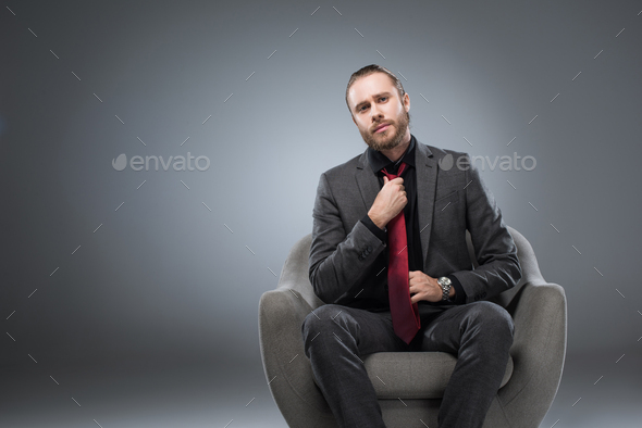 Serious businessman sitting in armchair and straightening tie, while looking at camera, isolated on