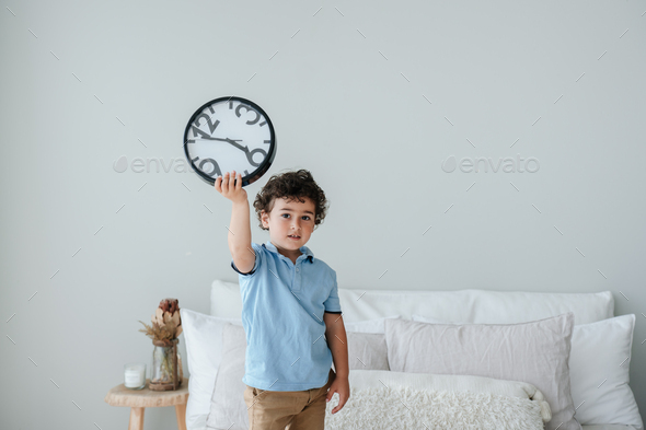 Cute caucasian boy in blue polo holding wall clock standing on bad, early morning getting up.