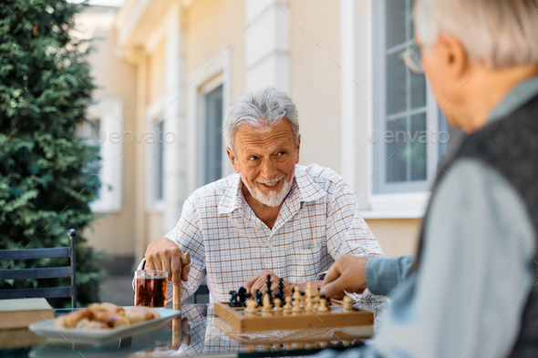 Happy senior man talking to his friend while playing chess on patio at retirement community.