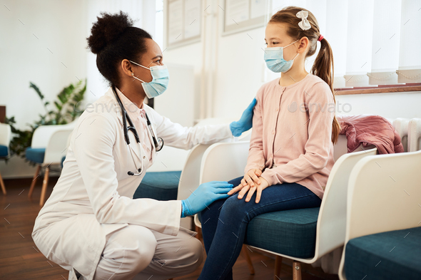 Little girl and black female doctor wearing face masks while talking in waiting room at clinic.