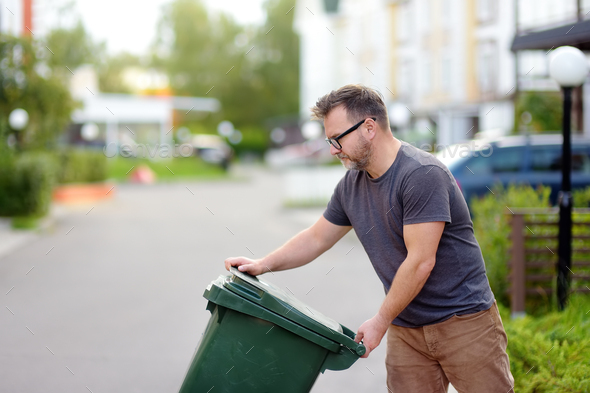 Mature man pulling out a large green plastic garbage container in front of the townhouse - Stock Photo - Images