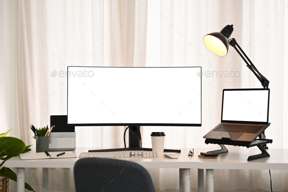 Ultra wide monitor, laptop, stationery and lamp on white table in modern creative office.