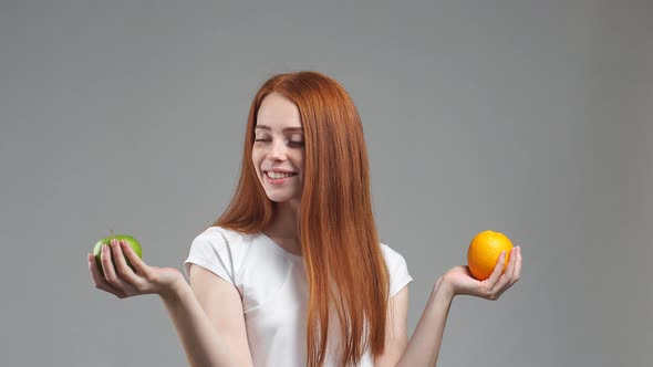 Beautiful Redhead Girl Tries To Choose Orange or Apple. Healthy Food Selection Concept.