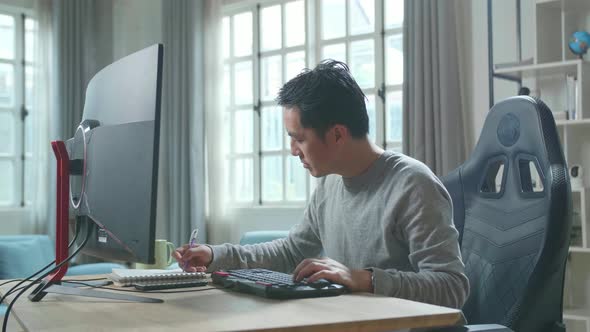 Asian Man Looking At Desktop Computer And Writing In Notebook While Working At Home alt