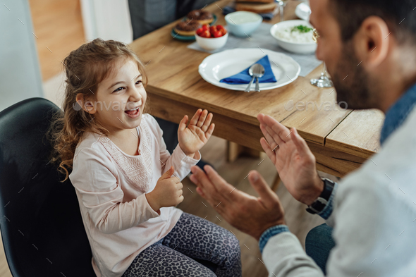 Happy little girl having fun while playing clapping game with her father in dining room.