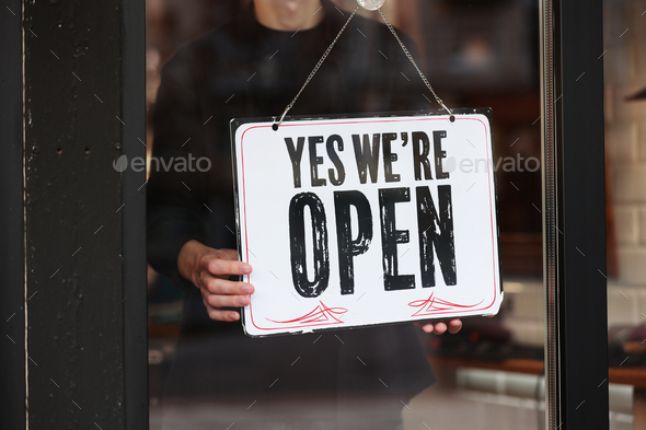 Man owner is hanging a chalkboard with inscription Yes we\'re open on glass door store, cafe, beauty