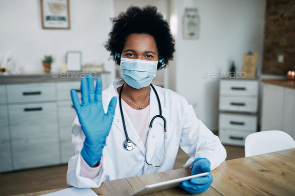 Black female doctor with protective face mask and glover using touchpad and waving to camera.