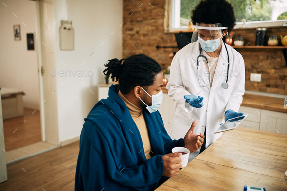 Black female doctor and her patient wearing face masks and using digital tablet during home visit.