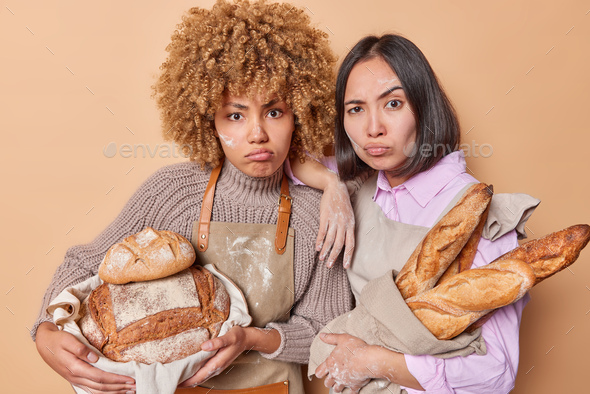 Serious attentive women bakers hold yeast free bread and baguettes focused displeased at camera wear