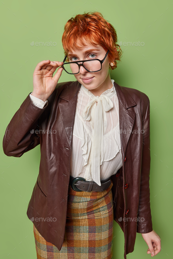 Vertical shot of serious intelligent female teacher keeps hand on rim of spectacles looks