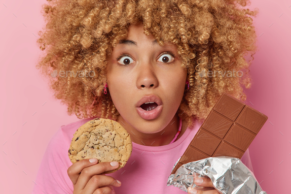 Close up shot of stunned woman holds breath from amazement keeps mouth opened holds cookie and bar
