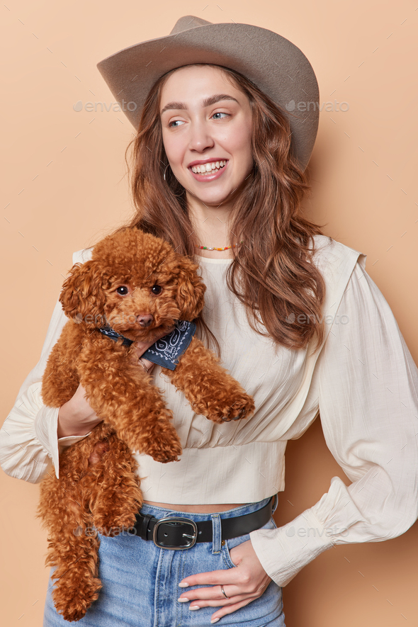 Vertical shot of cheerful woman wears cowboy hat blouse and jeans poses with small poodle puppy