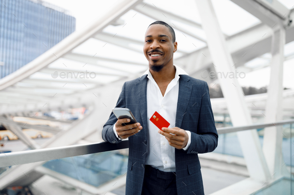 Black Businessman Buying Tickets Online With Smartphone And Credit Card At Airport