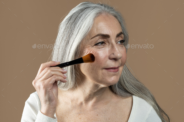 Glad old european lady with gray hair applies powder, blush with brush on her face