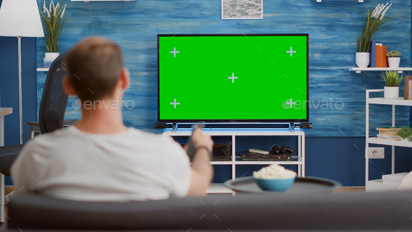 Man sitting on sofa looking at green screen on tv and switching channels