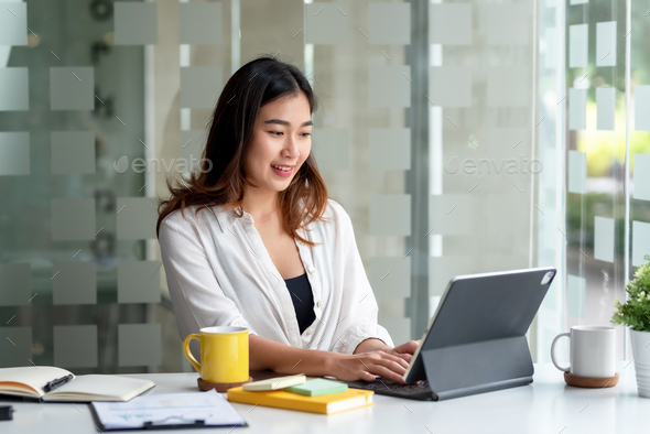 Attractive Asian business woman sitting at work happily working using tablet at office