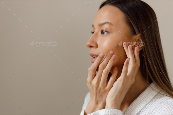 Upset young woman in white bathrobe examines pimples on her face. Portrait of girl removing pimples