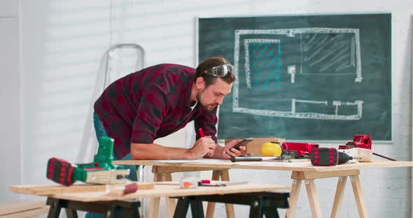 A Handsome Carpenter in a Flannel Shirt Counts the Dimensions on a Smartphone in a Carpentry alt
