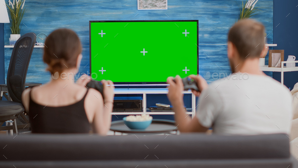 Couple holding controllers playing console online games on green screen tv sitting on couch