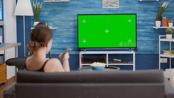 Young woman sitting on sofa looking at green screen on tv and switching channels
