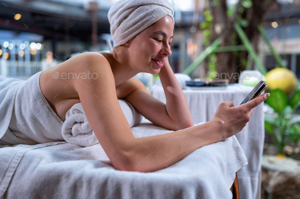 Young woman lying down on massage bed at wellness center check mobile phone social media network