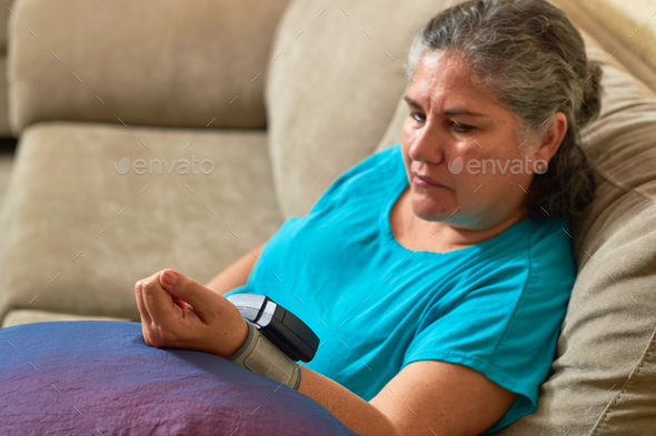 Mature Latina woman measures her blood pressure with digital monitor from home. Hypertension Concept