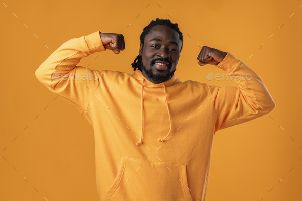 African smiling guy in yellow hoodie shows his physical strength against yellow background